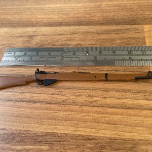 May include: A miniature brown rifle with black accents, approximately 12 inches long, is positioned next to a metal ruler. The ruler, marked in inches and millimeters, indicates the rifle's size. The rifle has a wooden stock and a metal barrel.
