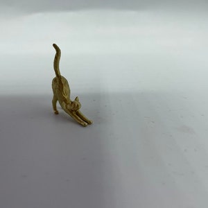 May include: A small, gold-colored metal figurine of a cat in a stretching pose. The cat is depicted with its front legs extended forward, its back arched, and its tail pointing straight up into the air, capturing a classic feline stretch.