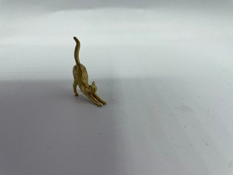 May include: A small, gold-colored metal figurine of a cat in a stretching pose. The cat is depicted with its front legs extended forward, its back arched, and its tail pointing straight up into the air, capturing a classic feline stretch.