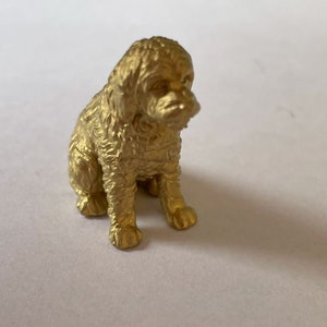 May include: A small, gold-coloured figurine of a dog sitting with its head tilted down. The dog has a fluffy, curly coat and is made of a smooth, shiny material.