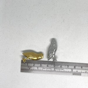 May include: Two small owl figurines, one gold and one silver, are placed on a white surface next to a ruler. The ruler is marked in inches and centimeters.