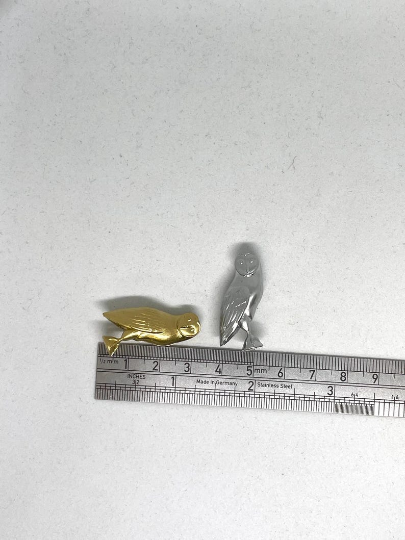 May include: Two small owl figurines, one gold and one silver, are placed on a white surface next to a ruler. The ruler is marked in inches and centimeters.