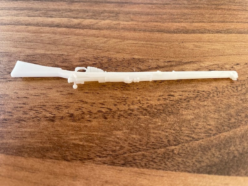 May include: A white, miniature rifle model, likely 3D-printed, with detailed features including a stock, barrel, and trigger guard. The toy rifle is set against a wooden background.