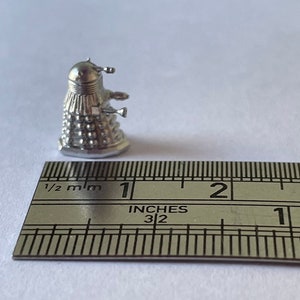 May include: A silver metal miniature of a Dalek, a fictional robot from the British science fiction television series Doctor Who. The Dalek is about 1 inch tall and is standing on a ruler.