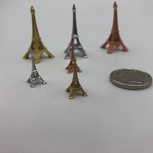 May include: Five miniature Eiffel Tower replicas in different sizes and colors: silver, gold, and rose gold. The towers are arranged on a white surface with a silver coin for scale.