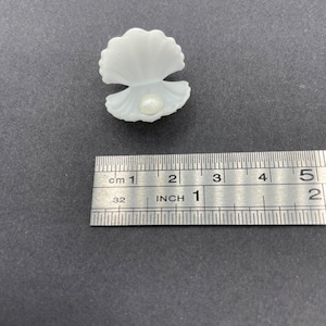 May include: A white plastic seashell with a pearl inside. The seashell is approximately 2.5 cm wide.