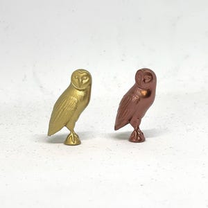 May include: Two small owl figurines, one gold and one copper, standing on a white surface.
