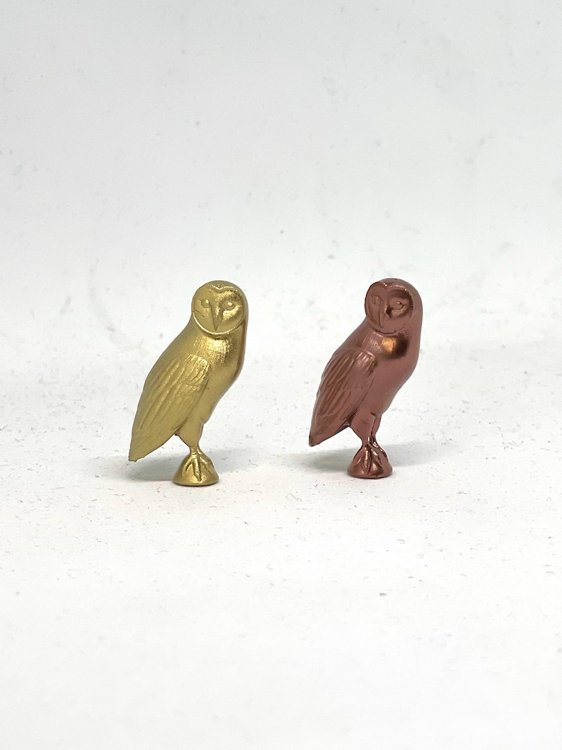 May include: Two small owl figurines, one gold and one copper, standing on a white surface.