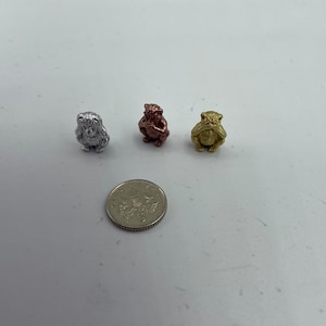 May include: Three small monkey figurines, each with a different hand gesture. The monkey on the left is silver, the middle monkey is copper, and the monkey on the right is gold.