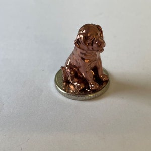 May include: A small, copper-colored figurine of a dog sitting on a silver coin. The dog is facing to the right and has a detailed, realistic appearance.