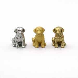 May include: Three small dog figurines in silver, gold, and brass tones are arranged in a row. The dogs are sitting, with their front paws extended. The background is plain white.