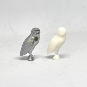 May include: Two owl figurines, one silver and one white, are shown on a white background. The owls are facing forward with their wings folded.