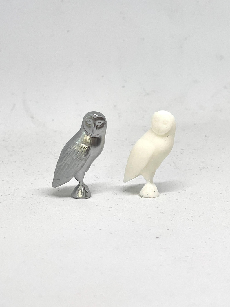 May include: Two owl figurines, one silver and one white, are shown on a white background. The owls are facing forward with their wings folded.