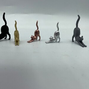 May include: Five small cat figurines in a stretching pose, arranged in a row. The cats are finished in matte black, gold, copper, silver, and dark gray metallic tones. Each figurine features an arched back, extended front paws, and a raised tail.
