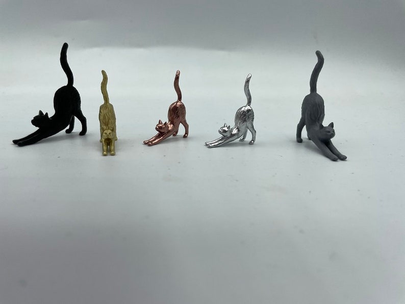 May include: Five small cat figurines in a stretching pose, arranged in a row. The cats are finished in matte black, gold, copper, silver, and dark gray metallic tones. Each figurine features an arched back, extended front paws, and a raised tail.