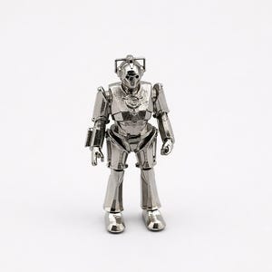 May include: A silver-colored Cyberman action figure, a fictional cyborg from the Doctor Who series, stands upright against a white background. The figure has a detailed metallic design, including a helmet, chest plate, and articulated limbs.