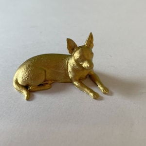 May include: A gold-colored figurine of a small dog lying down. The dog has floppy ears and a short tail.