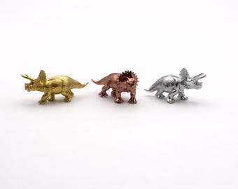 May include: Three small triceratops figurines in gold, copper, and silver. The dinosaur toys are displayed on a white background. Each figurine has a distinct metallic finish, showcasing the details of the prehistoric creature.