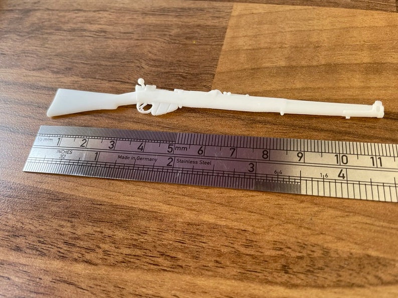 May include: A white plastic toy rifle, approximately 11 inches long, is positioned next to a metal ruler. The ruler displays both inches and millimeters. The rifle has detailed features and is set against a wooden surface.