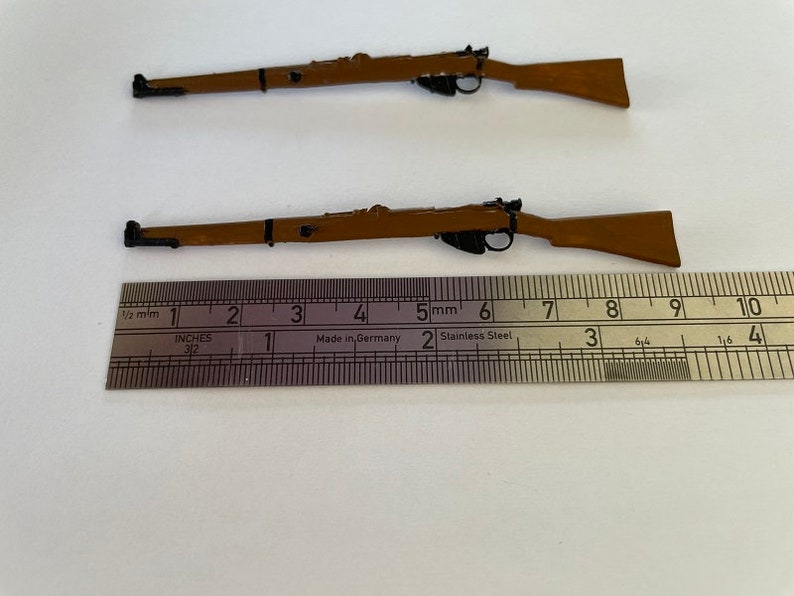 May include: Two miniature brown rifles with black details are displayed. A ruler with inch measurements is placed below the rifles. The rifles appear to be replicas or models, possibly for collectors or hobbyists.