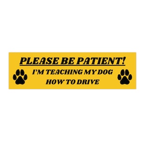 May include: Yellow rectangular bumper sticker with black text: "PLEASE BE PATIENT! I'M TEACHING MY DOG HOW TO DRIVE." Black paw prints are on each side of the text.