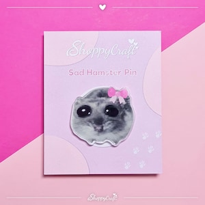 Könnte beinhalten: Anstecknadel mit einem traurigen Hamster mit großen schwarzen Augen und einer rosa Schleife. Die Anstecknadel befindet sich auf einer rosa Karte mit dem Text "ShoppyCraft" und "Sad Hamster Pin". Der Hintergrund ist ein Rosé-Farbverlauf.