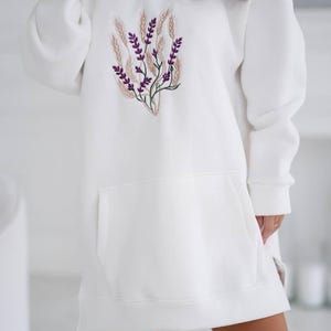 May include: White oversized hoodie with a floral embroidery design featuring purple flowers and beige stems. The hoodie has a kangaroo pocket and a drawstring hood.