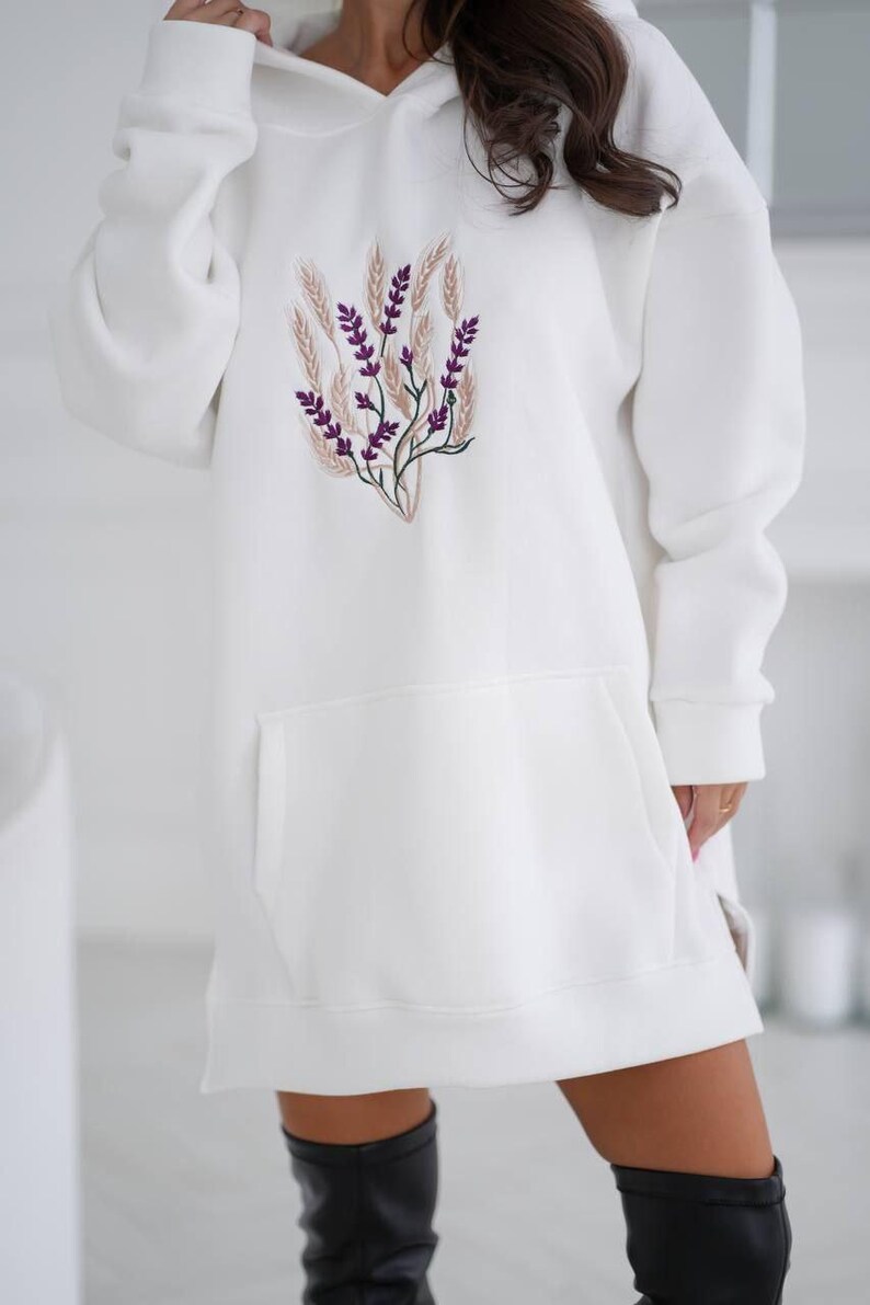 May include: White oversized hoodie with a floral embroidery design featuring purple flowers and beige stems. The hoodie has a kangaroo pocket and a drawstring hood.