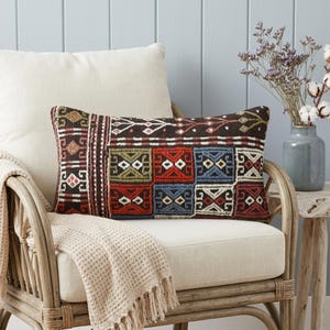 Puede incluir: Un cojín rectangular con una base de color marrón oscuro y un patrón geométrico multicolor en rojo, azul, verde y blanco. El diseño consta de motivos cuadrados repetitivos y bordes lineales, mostrando una apariencia tejida y texturizada.