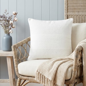 Pode incluir: Uma almofada quadrada branca texturizada repousa sobre uma cadeira de vime com uma almofada creme. Uma manta bege com textura de waffle e franjas está sobre a cadeira. Um vaso azul com flores secas está sobre uma mesa lateral de madeira, contra uma parede azul clara.