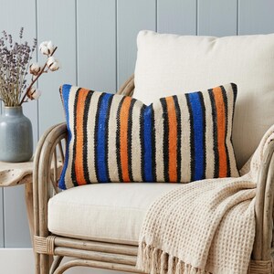 May include: A rectangular throw cushion featuring a vertical striped pattern in vibrant orange, deep blue, and cream colours. The fabric has a textured, woven appearance with consistent, repeating lines across the entire surface of the cushion.
