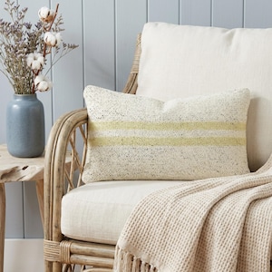May include: A rectangular decorative pillow with a cream-colored speckled fabric and two horizontal yellow stripes. The pillow is on a cream-colored chair with a waffle-knit throw blanket. A blue vase with dried flowers sits on a wooden side table.