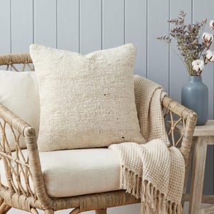 以下が含まれることがあります： クリーム色のテクスチャード加工のクッションが、お揃いのクッションが付いた籐の椅子に置かれています。フリンジ付きのワッフル織りのスローが椅子に掛けられています。ドライフラワーと綿の茎が入った花瓶が小さな木製のテーブルに置かれています。