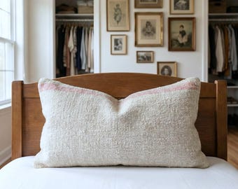 Puede incluir: Una almohada rectangular blanquecina con una superficie texturizada y una franja rosa en el borde superior descansa sobre una estructura de cama de madera. La cama está cubierta con una sábana blanca. La habitación presenta obras de arte enmarcadas.