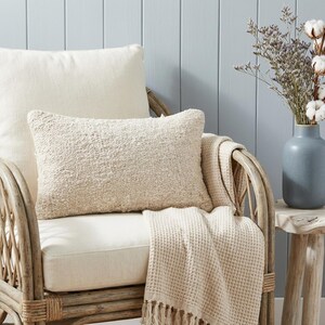 May include: A cream-colored textured lumbar pillow rests on a wicker chair, alongside a matching throw blanket with fringe. A second, larger pillow is behind it. A blue vase with dried flowers sits on a small wooden side table.