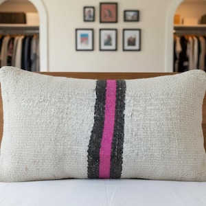Peut inclure: Un coussin rectangulaire texturé, de couleur crème. Une bande verticale noire et une bande rose vif traversent le centre. Le coussin est posé sur une surface blanche, avec une tête de lit en bois en arrière-plan.