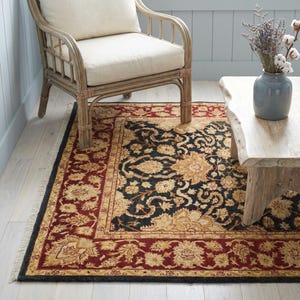 Puede incluir: Una alfombra rectangular con un diseño floral tradicional. La alfombra presenta un centro negro con patrones florales dorados, un borde burdeos y un fleco negro. Una silla de mimbre de color claro y una mesa de madera están en el fondo.