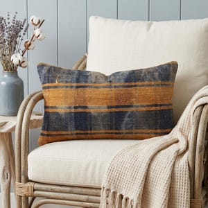 May include: A rectangular throw pillow featuring a woven plaid pattern in shades of dark navy blue and mustard yellow. The fabric has a visible, textured weave, and the pillow is positioned on a light-colored chair with a matching cream-colored cushion.