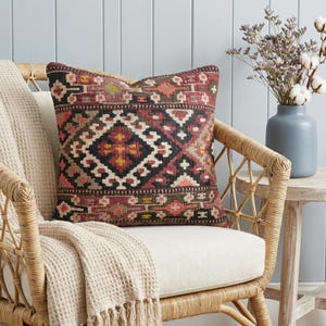 May include: A patterned cushion in burgundy, black, cream, and gold tones, placed on a wicker chair with a cream blanket. The cushion features a geometric design. A vase of dried flowers sits on a small wooden table nearby.