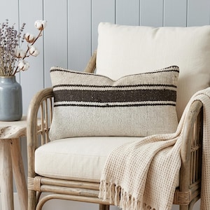 May include: A rectangular cushion with three dark brown stripes rests on a cream-coloured chair. A cream-coloured blanket with a fringe is draped over the chair. A vase of lavender and cotton stems sits on a wooden table.