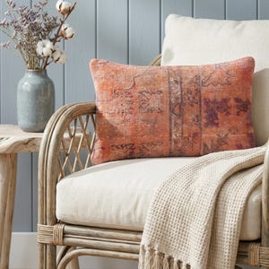 Peut inclure: Un coussin rectangulaire à motifs orange et violet repose sur une chaise crème. Le coussin a un aspect vieilli et vintage. Un plaid crème en nid d'abeille avec des franges est drapé sur la chaise. Un vase avec des fleurs séchées est à proximité.