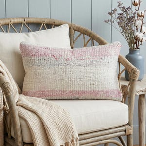 Puede incluir: Un cojín rectangular de tela tejida con textura y rayas horizontales en tonos crema, rosa suave y gris apagado. El cojín presenta un patrón de tejido grueso visible y está colocado sobre el cojín de una silla de color claro.