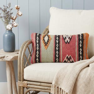 Puede incluir: Cojín decorativo rectangular con un diseño tejido en tonos rojos, naranjas, negros y crema. El cojín está colocado sobre una silla color crema con estructura de ratán. Una manta color crema con flecos está sobre la silla. Mide aproximadamente 40 cm x 20 cm.