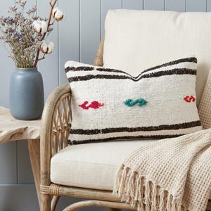 May include: A rectangular white pillow with black stripes and colorful embroidered accents rests on a cream-colored chair. The pillow has red and teal embroidered details. A cream-colored waffle-weave throw with fringe is draped over the chair.