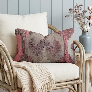 May include: A decorative lumbar pillow with a geometric pattern in shades of pink, gray, and burgundy rests on a cream-colored chair. A cream-colored throw blanket with fringe is draped over the chair. A vase of dried flowers sits on a small wooden table.