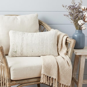 Può includere: Un cuscino rettangolare color crema con una superficie strutturata poggia su una sedia in vimini con un cuscino quadrato abbinato. Una coperta a nido d'ape con frange e un vaso blu con fiori secchi completano la scena.