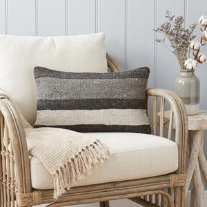 May include: A striped cushion in shades of grey and cream, placed on a cream-coloured chair with a rattan frame. The cushion has a rectangular shape. A cream throw with fringing is draped over the chair. The scene evokes a relaxed, neutral aesthetic.