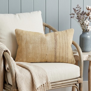 May include: A rectangular lumbar pillow featuring a textured, woven fabric in a light tan or beige hue. The pillow displays a subtle, vertical dark brown stripe pattern running along the right side, set against a neutral, coarse-weave textile.