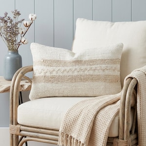 Puede incluir: Un cojín rectangular color crema con rayas horizontales color beige y detalles texturizados descansa sobre una silla color crema. Una manta a juego con flecos está sobre la silla. Un jarrón con flores secas está cerca.