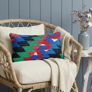 May include: A rectangular decorative pillow with a geometric pattern in red, blue, green, and black. The pillow is placed on a cream-colored cushion inside a wicker chair. A cream-colored throw blanket with fringe is draped over the chair.
